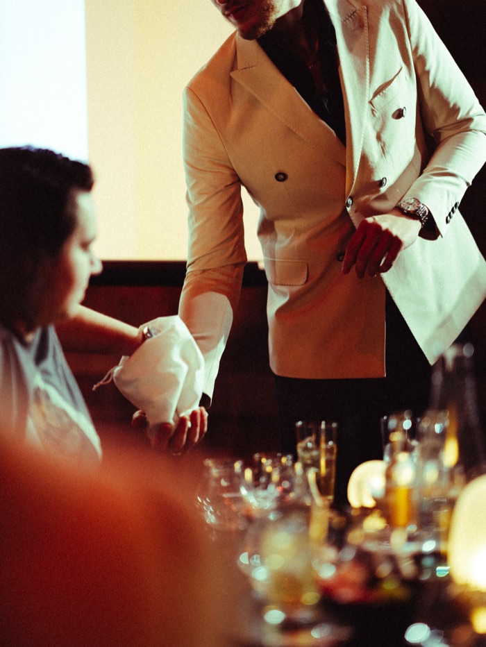 A sensory task at a Sense Shift tasting: a guest's hand inside a cloth bag, the host in a white jacket at a table full of glasses